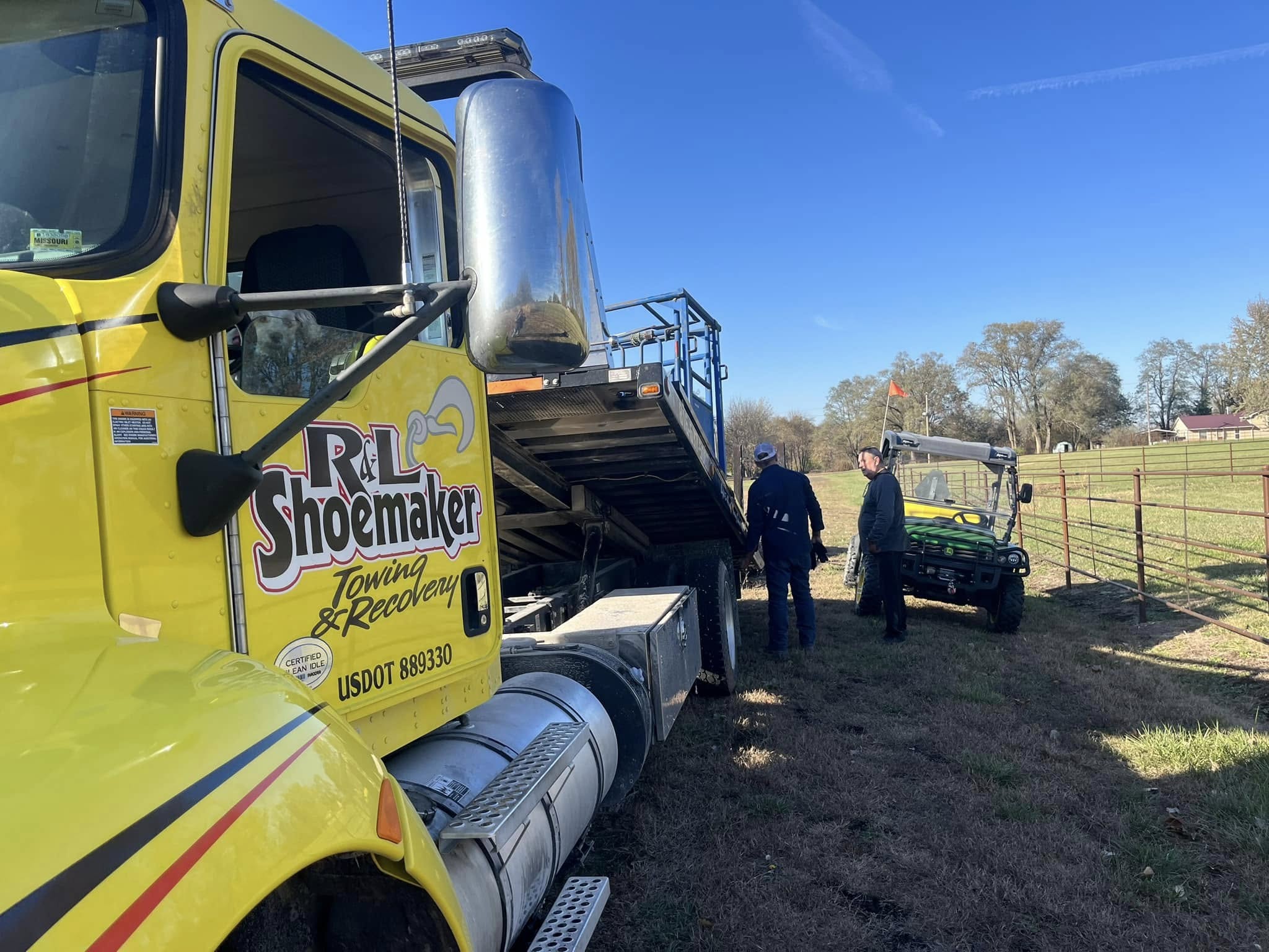 Yellow tow truck beside a tilted flatbed trailer on a grassy field. Two people stand near an off-road vehicle. Clear blue sky and trees in the background.